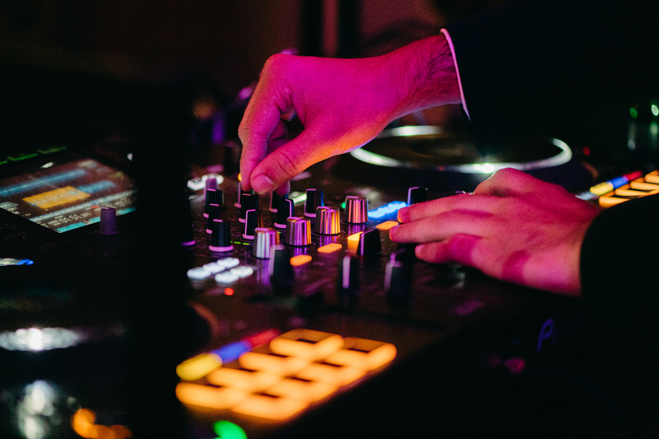 Close-up of a DJ's hands adjusting sound levels on a mixer, bathed in moody purple and amber lighting during a wedding celebration.