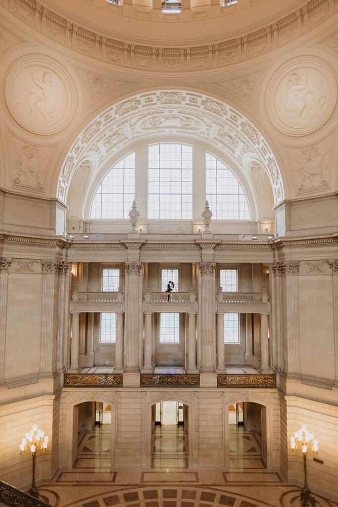 A couple sharing a romantic moment on the upper balcony of San Francisco City Hall, framed by massive arched windows and sweeping classical columns beneath a domed ceiling.