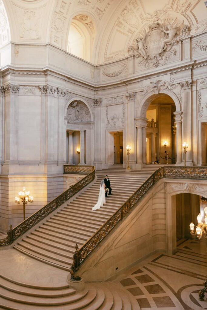 Bride and groom captured on the grand staircase of San Francisco City Hall, surrounded by soaring Beaux-Arts architecture, ornate gold detailing, and soft amber lighting.