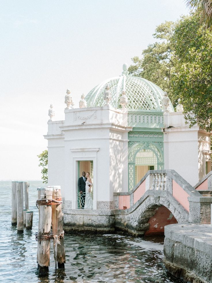 A bride and groom framed in the arched doorway of Vizcaya’s pastel green lattice pavilion, perched at the water’s edge with a romantic European coastal feel.