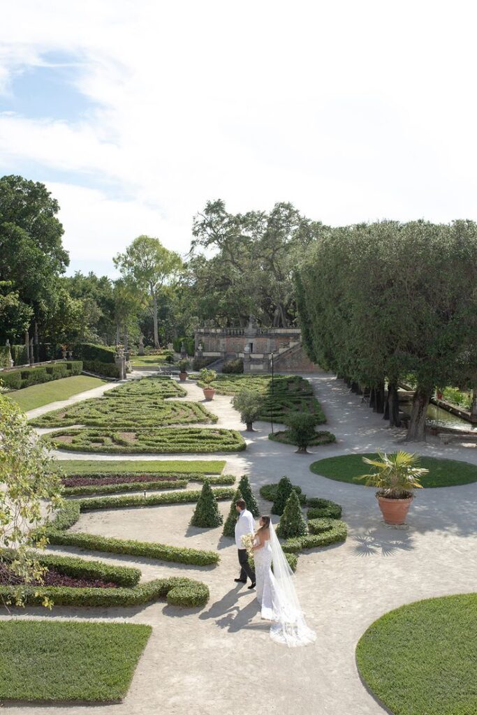 A bride and groom walk hand-in-hand through Vizcaya’s manicured gardens, surrounded by sculpted hedges and sun-drenched paths in a lush, elegant setting.