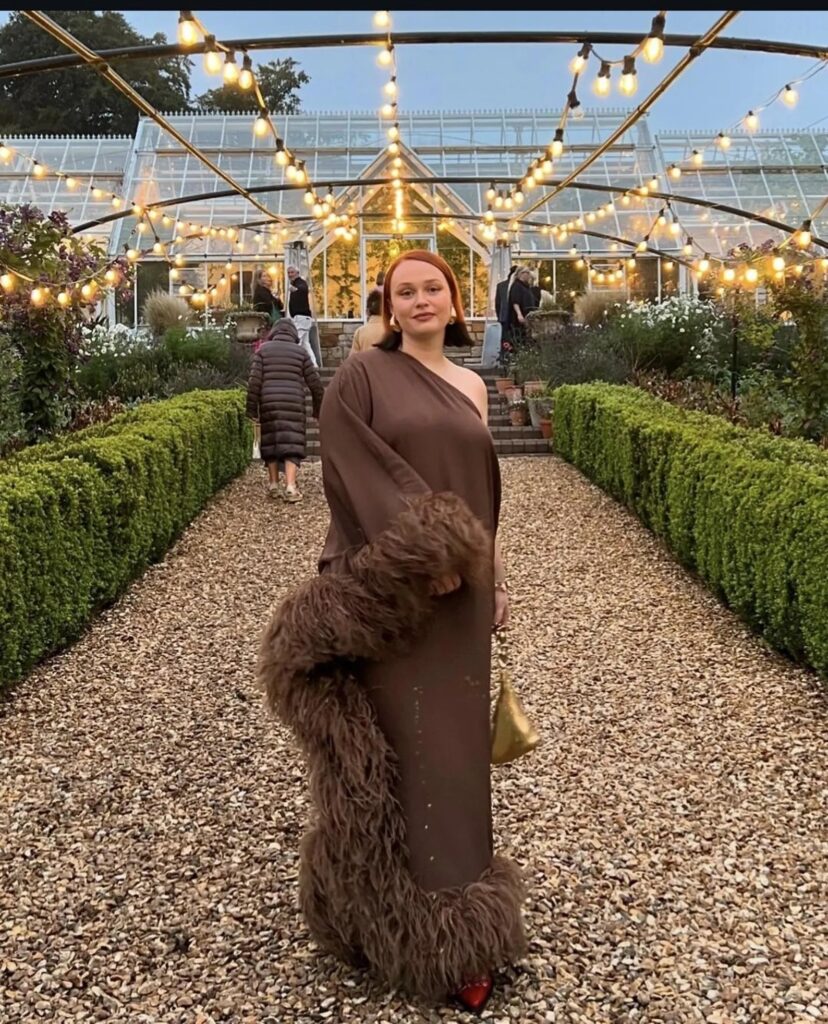 Wedding guest in a dark brown one-shoulder gown with feather trim and sculptural draping — dramatic and fashion-forward guest attire for a moody, editorial wedding.